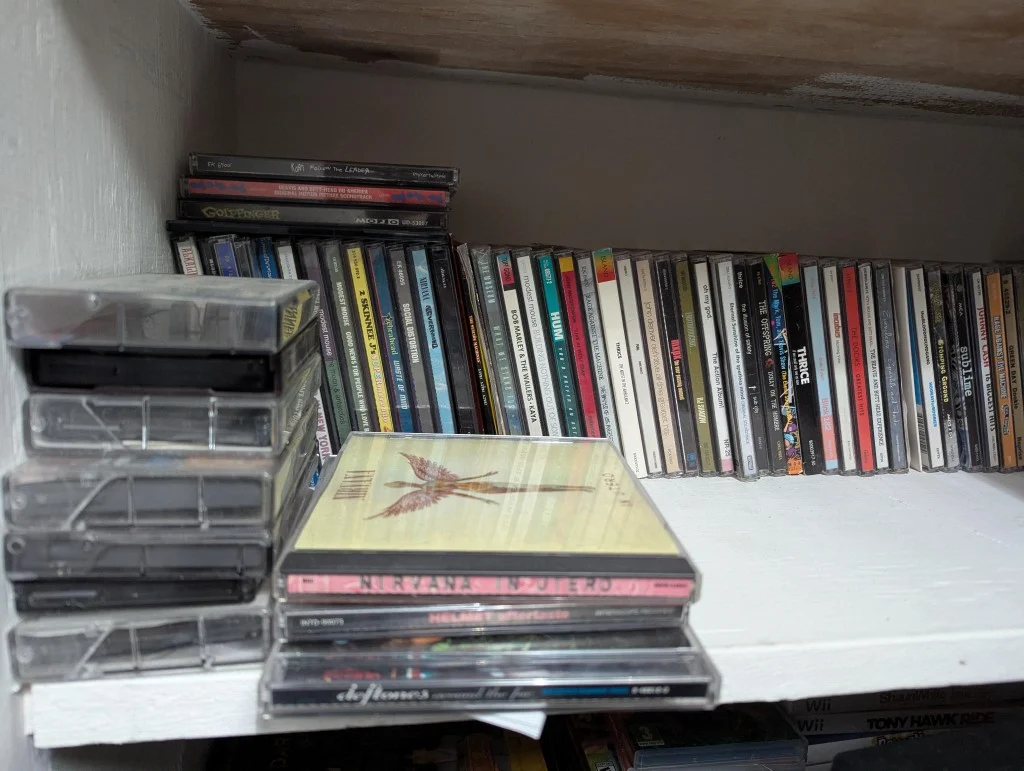 A white wooden shelf with a stack of three CD jewel cases in the foreground, a row of album spines behind them, and a column of cassette tapes in clear cases on the left—personal music media collection.