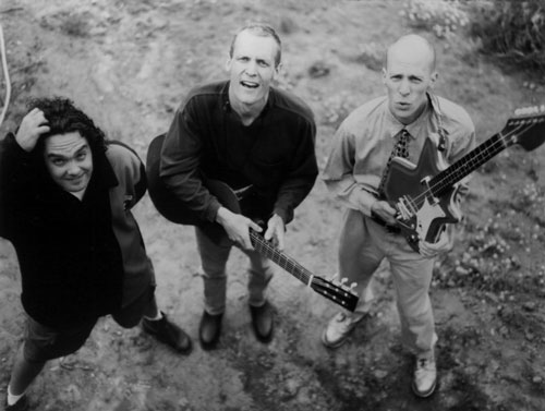 Black-and-white high-angle promotional photograph of the Presidents of the United States of America on bare ground—drummer Jason Finn at left standing by a small kit, Dave Dederer at center holding a guitbass, Chris Ballew at right holding a basitar—all looking up toward the camera.